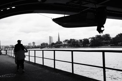 Black and white street photo of two figures under a riverside overpass in Frankfurt, with the River Main, ECB building, and Dreikönigskirche spire in view.