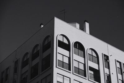 Black and white photo of a modernist apartment corner with arched windows, balcony grids, sunlit white brick, and deep shadow against a clear sky.
