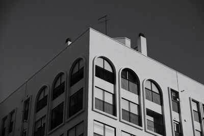 Black and white photo of a modernist apartment corner with arched windows, balcony grids, sunlit white brick, and deep shadow against a clear sky.